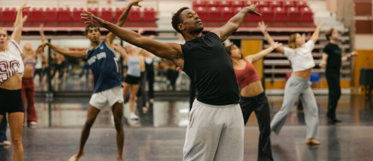 Marquez dances in front of students in a studio. Wearing sweatpants and a tank top, he leans back with his arms extended above his head.