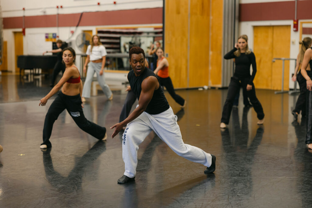 Marquez dances alongside students in a dance studio. He lunges with his arm across his body. He wears sweatpants, a tank top, and socks.
