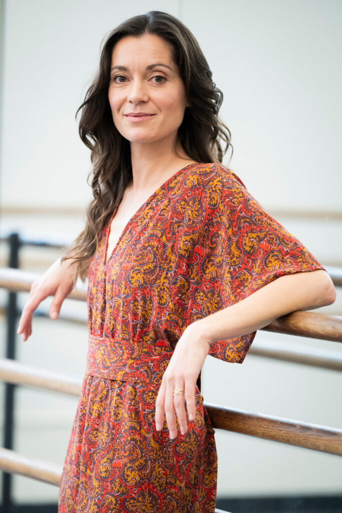 Melissa Toogood wears a pink and brown dress and leans against the barre in a dance studio, smiling at the camera.