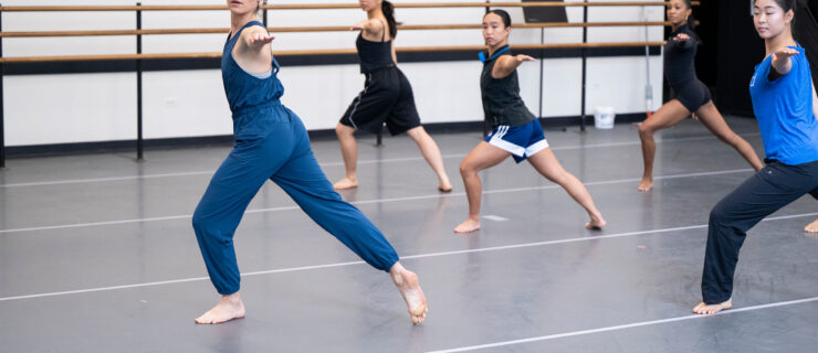 Melissa Toogood wears a blue jump suit as she lunges in front of a class of dancers copying her movement. They are in a dance studio with gray marley flooring and a barre along the wall.