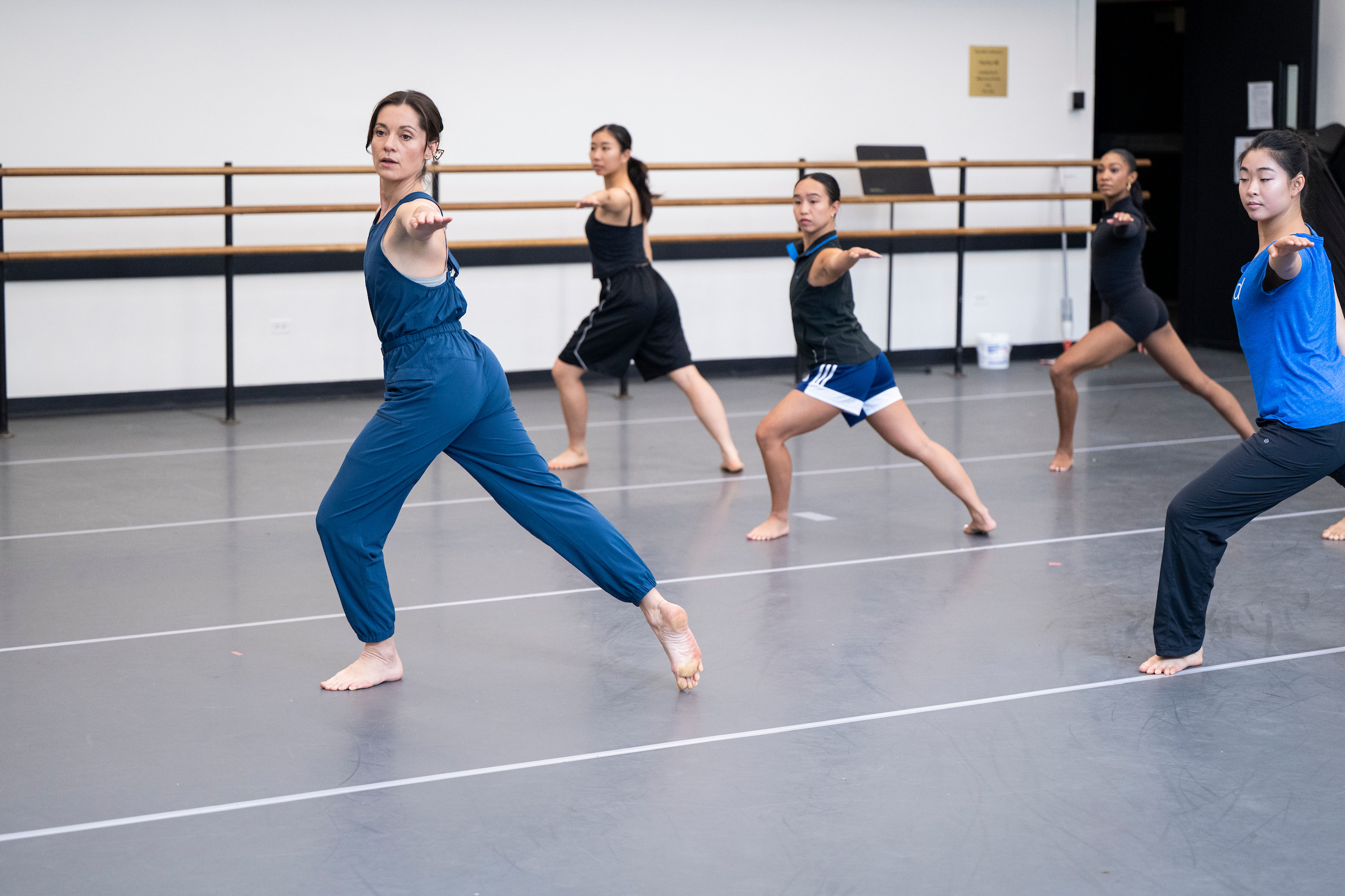 Melissa Toogood wears a blue jump suit as she lunges in front of a class of dancers copying her movement. They are in a dance studio with gray marley flooring and a barre along the wall.