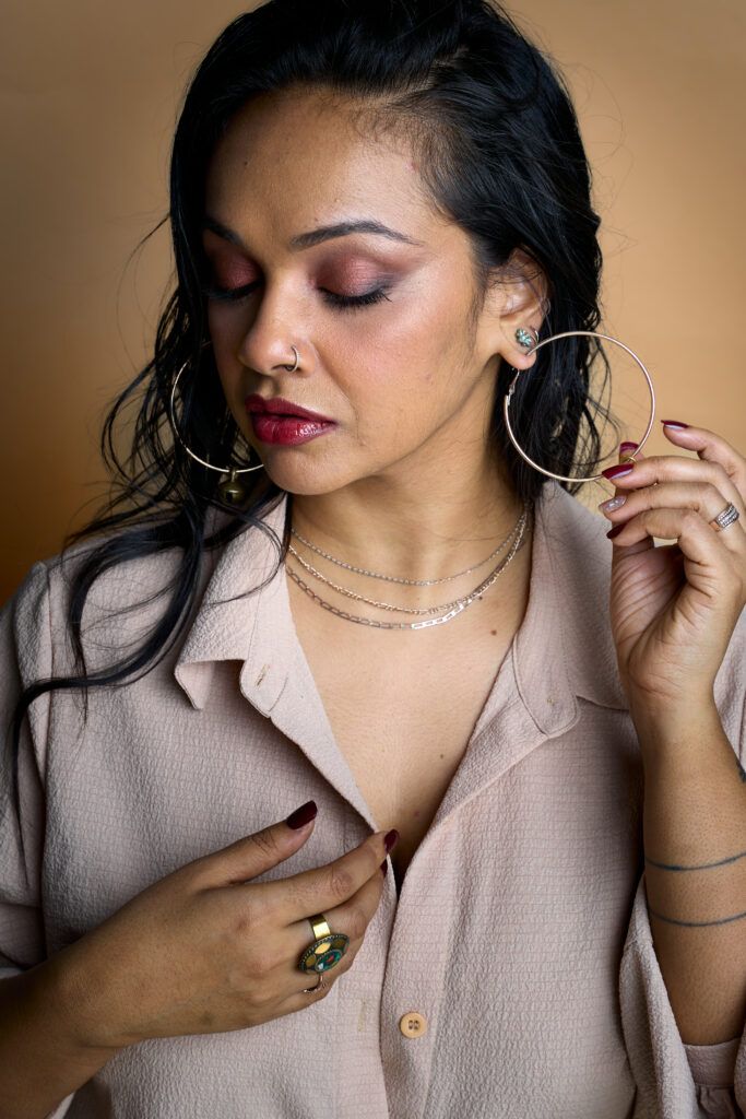 Brinda Guha poses holding one hoop earring with her eyes downcast. She wears a cream blouse against a tan background.