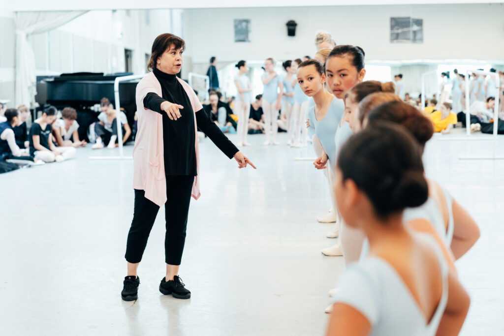 In a ballet studio, Gabay speaks to young dancers wearing light blue leotards and tights. They stand in a long line as they listen.