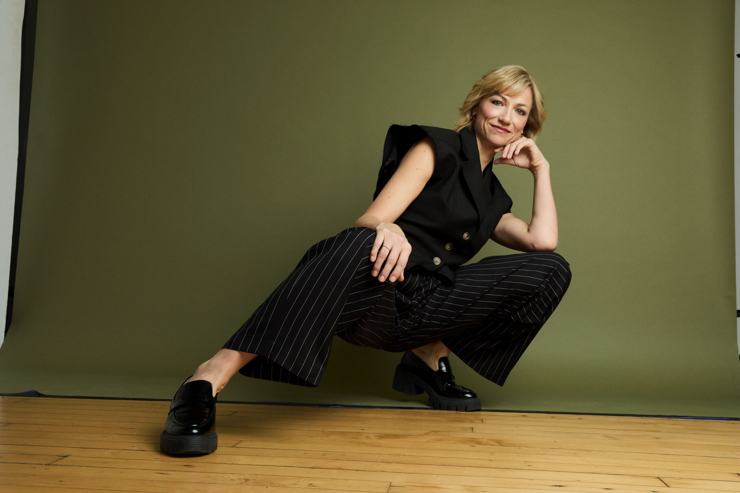 Tap artist Sara Brians squats towards the left with one leg extended out to the right. Her left arm is propped on her chin. She is dressed in a black outfit and black shoes.